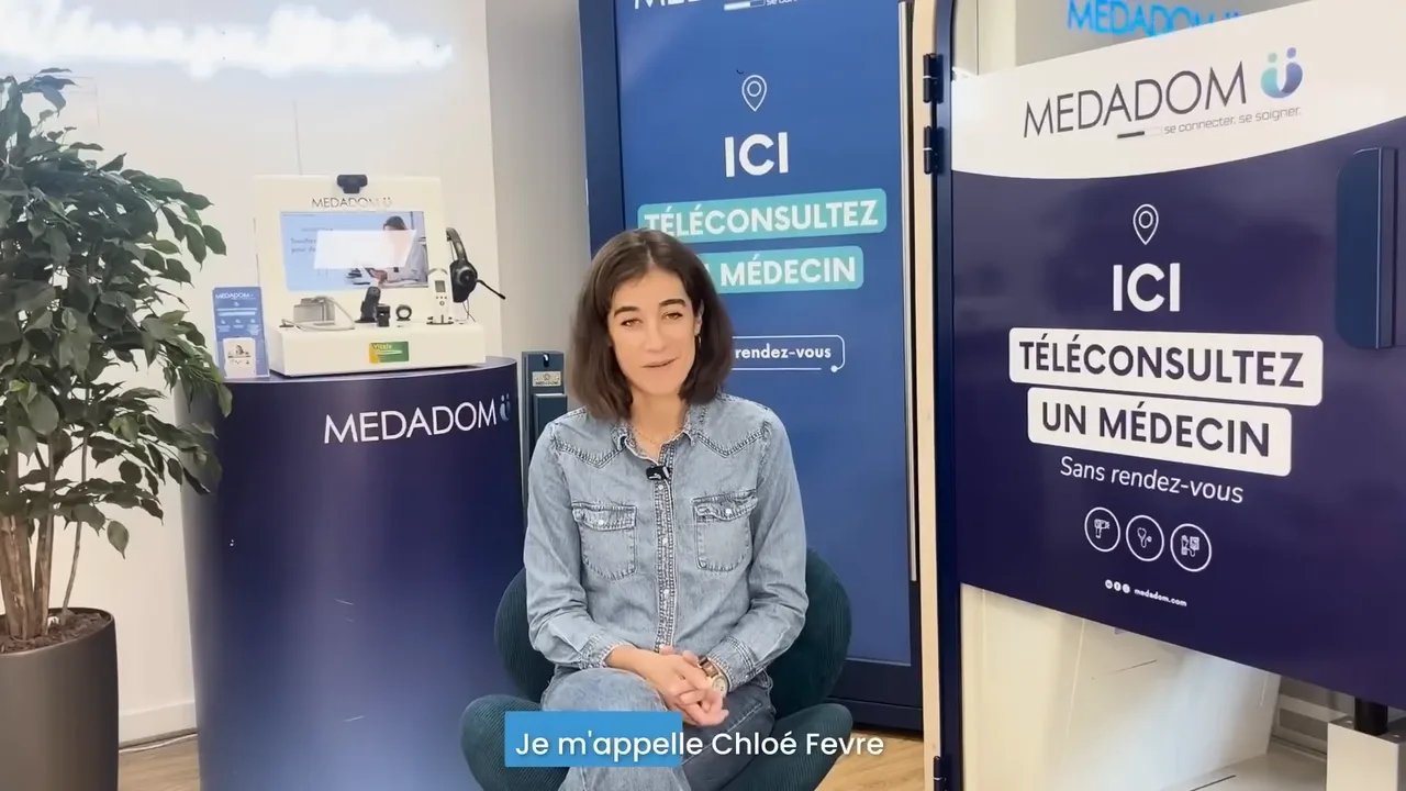 Dr Chloé Fevre, assise entre une borne de téléconsultation MEDADOM et un panneau « Ici, téléconsultez un médecin, sans rendez-vous », se présente face caméra