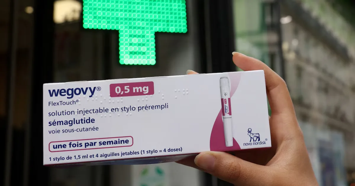 Main tenant une boîte de Wegovy devant une croix verte de pharmacie